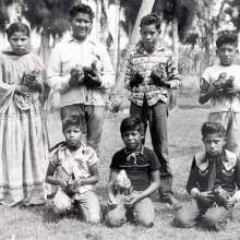  children lined up outside holding chickens. The chickens were part of a project at the Brighton Indian Day School. Standing in the back are Elsie Jean Bowers, Stanley Huff, Coleman Josh, and unidentified (Mr. Boehmer listed this person as unidentified, but several Tribal members have identified him as Fred Smith). Kneeling in the front are Eddie Shore, Jommy Scott Osceola, and Jerry Micco.