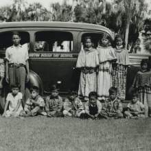  students at the Brighton Indian Day School gathered around a vehicle (most likely the car that brings the children to school). Standing from left to right are Jack Micco, Harry Tommie, Storeman Osceola, Maude Johns, Lottie Johns, Lauderdale Johns, Mary Jumper, Louise Smith (seated on fender). Seated from left to right are Wonder Johns, Howard Micco, Joe Jumper, unidentified, Archie Johns, Cecil Johns, unidentified.