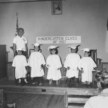  graduating kindergarten class of 1957 in their caps and gowns sitting on a stage. The children are identified as Judy Jones, Willie Johns, Patty Johns, Frank Huff, Jr., and Loma Norman Johns. Standing behind Jody Jones is the children's teacher Edmund Harjo.